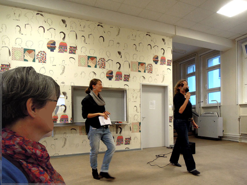 Eine Schulmensa wird gezeigt mit Essenausgabe. An der Wand ist eine Tapete aus Kinderzeichnungen zu sehen. Sie besteht aus Linien von gezeichneten Köpfen und mit farbigem Buntstiften gefüllten Kopfprofilen. Vor der Tapete sind drei Erwachsene der Schulleitung zu shen, die die nach mit den lernenden Kindern durchgeführten Kunstprojekt von Anett Lau eröffnen.